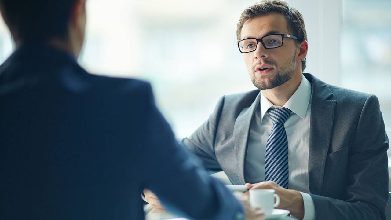 Imagem mostra dois homens de terno conversando. No primeiro plano, aparece um deles de costas, conversando com o outro, que está de frente.