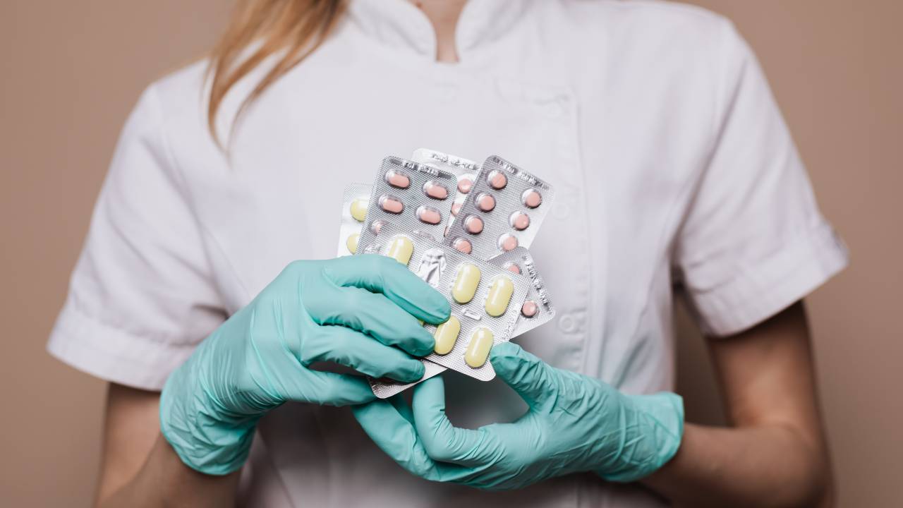 Imagem de uma com roupas hospitalares, usando luvas e segurando embalagens de remédio.
