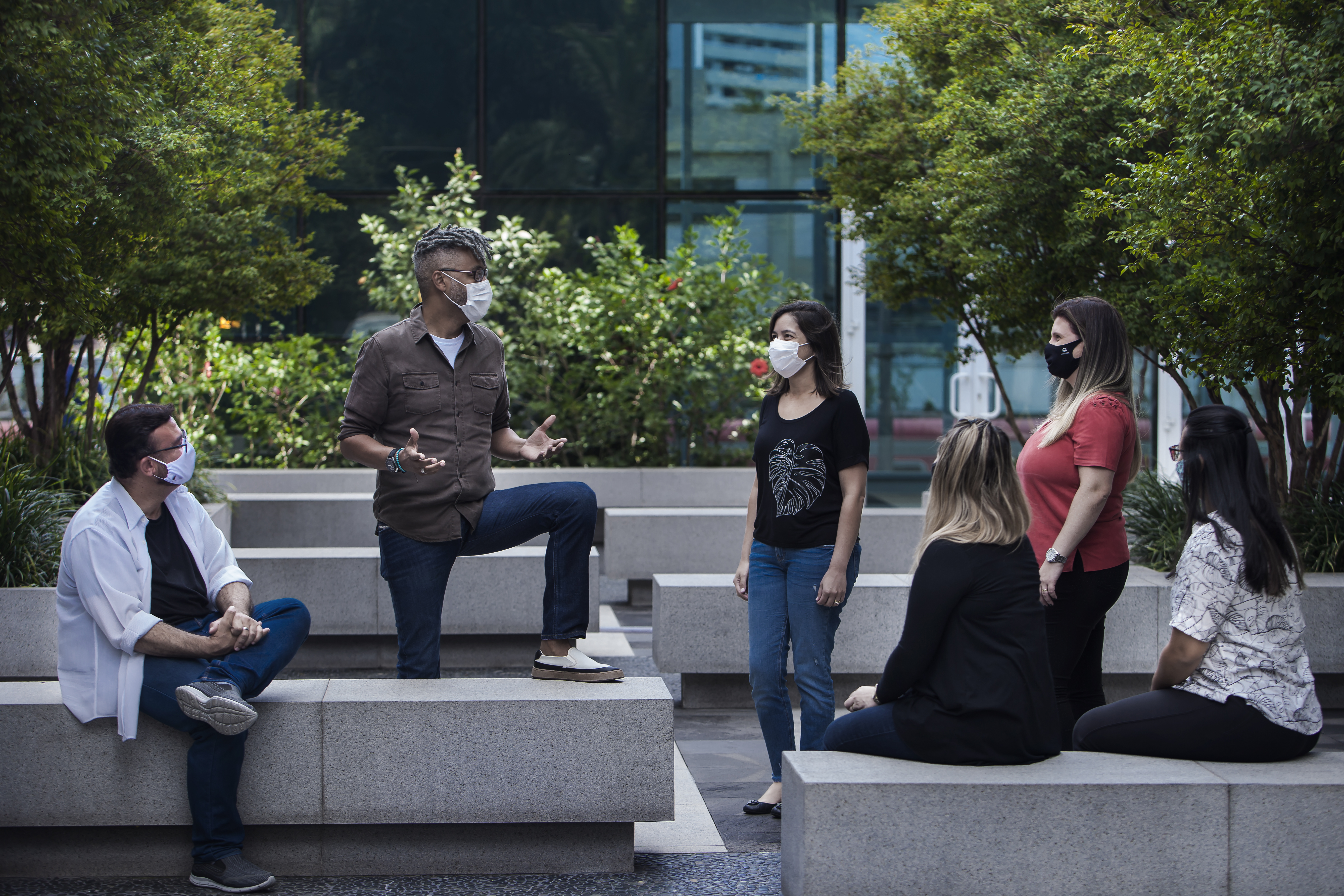 Imagem mostra um grupo de 5 pessoas conversando, usando máscaras, em um jardim. Ao centro está Moisés Marques, um homem negro de camisa marrom.