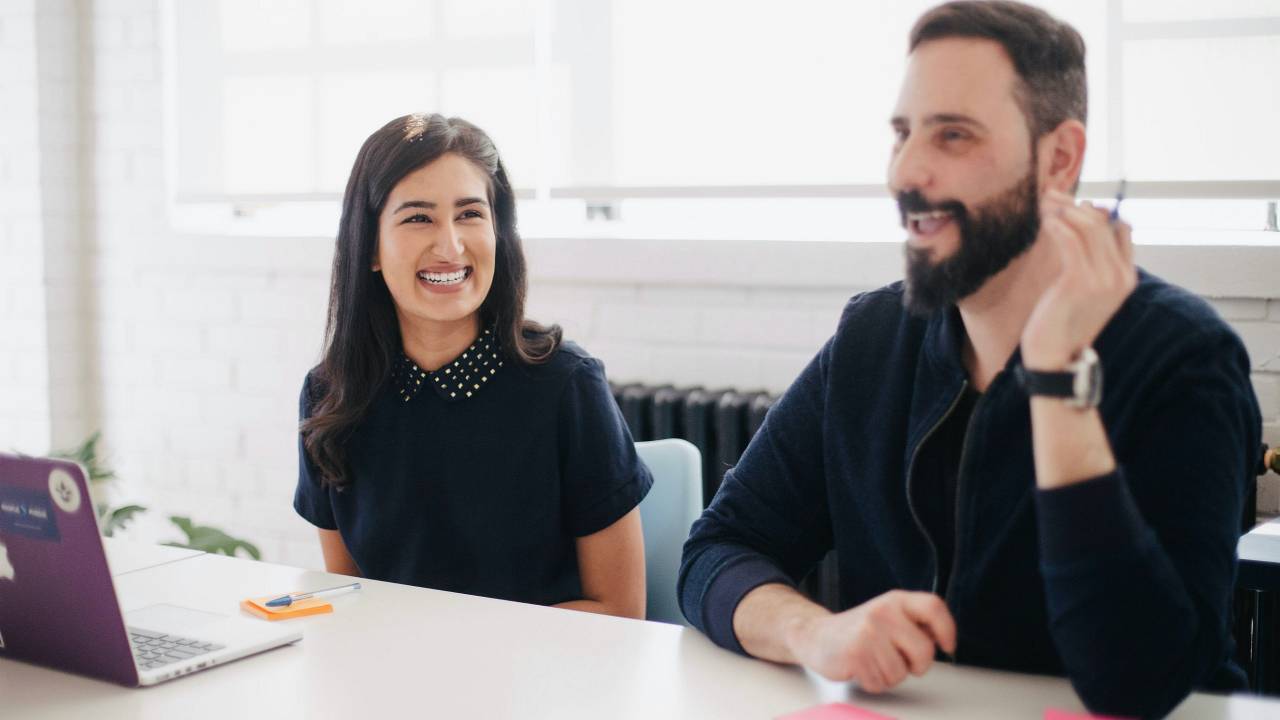 Imagem mostra uma mulher e um homem sentados em uma mesa de reunião. Ele está falando algo e ela está rindo