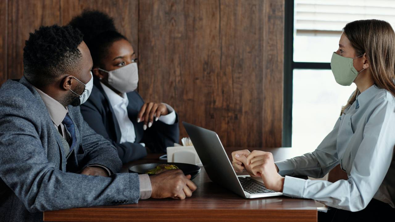 Imagem mostra três pessoas em uma mesa de reunião, usando máscaras: um homem negro, uma mulher negra e uma mulher branca, que está em frente a um notebook.