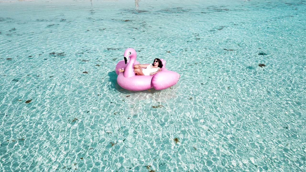 Imagem mostra uma mulher de maiô branco em cima de uma boia rosa em formato de Flamingo no meio de um mar azul claro.