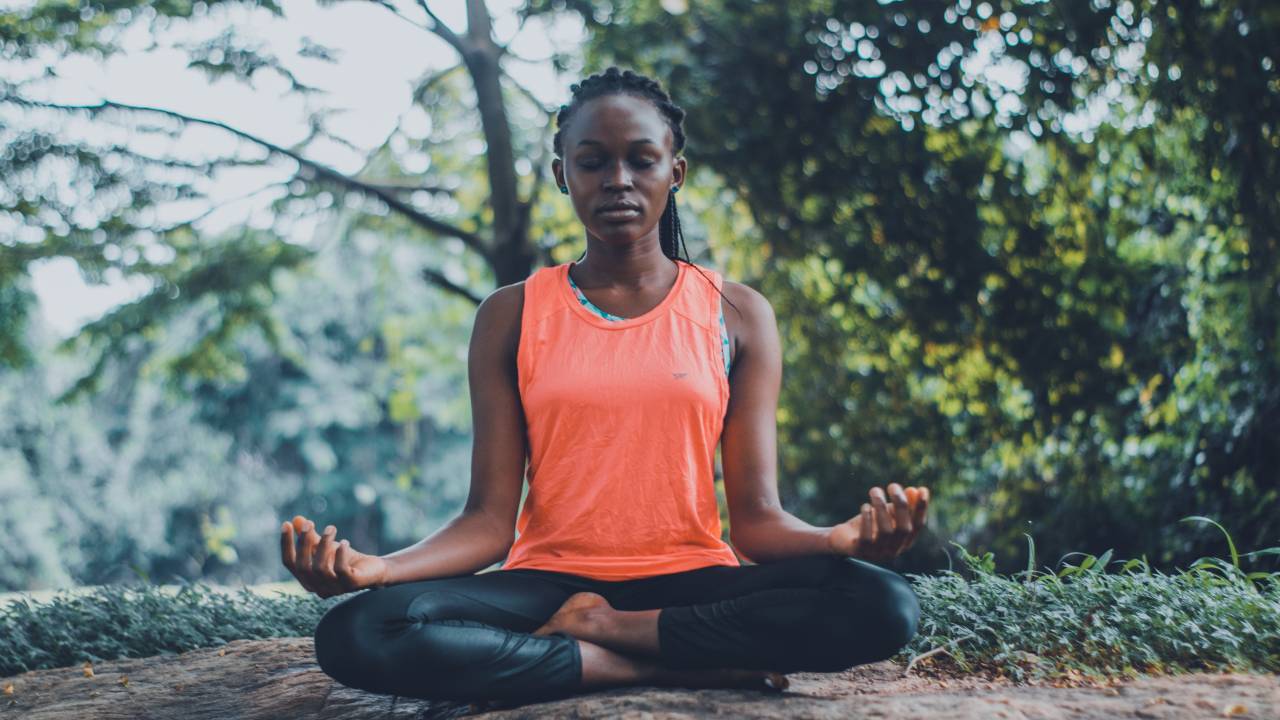Imagem mostra uma mulher negra de cabelos presos e trançados sentada em posição de meditação em uma área verde.