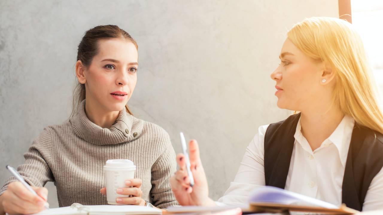 Imagem mostra duas mulheres brancas (uma de cabelos castanhos e outra de cabelos loiro) conversando em uma mesa de reunião.