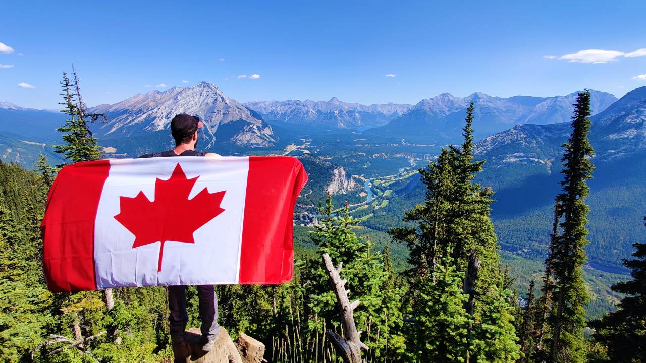 Imagem mostra um homem de costas segurando uma bandeira do Canadá. Ele está no alto de uma montanha.