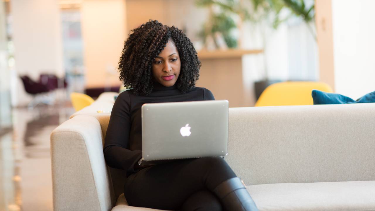 Imagem mostra mulher negra, sentada em um sofá, usando um notebook.