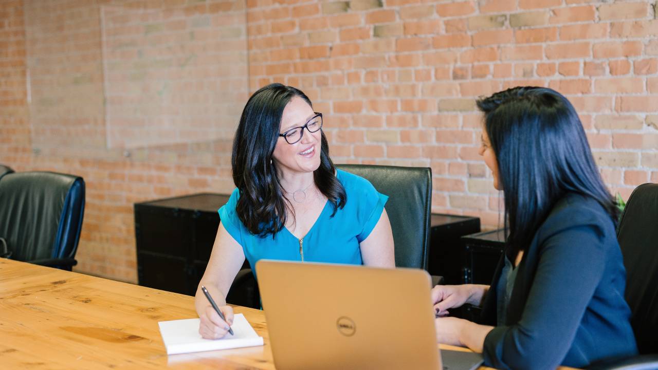 Foto mostra duas mulheres brancas sentadas lado a lado numa mesa de trabalho, conversando