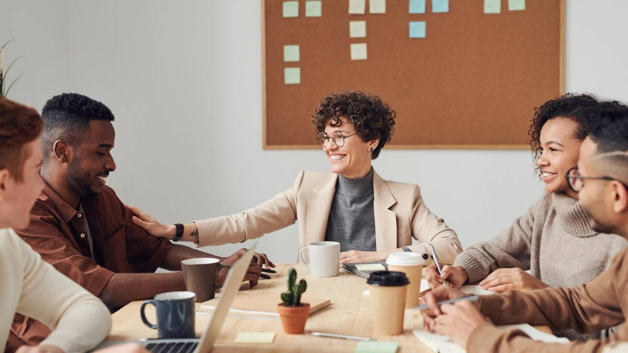 Imagem mostra um grupo de pessoas sentado em uma mesa de reunião. Em primeiro plano, uma mulher negra de cabelos curtos e cacheados coloca a mão no braço de um homem negro. Todos estão sorrindo.