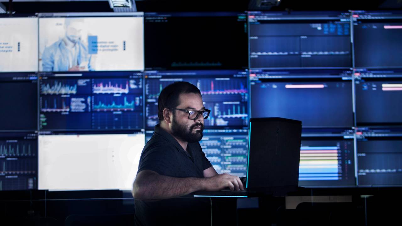 A imagem mostra Marcus Garcia mexendo no computador em um ambiente cheio de telas azuladas.