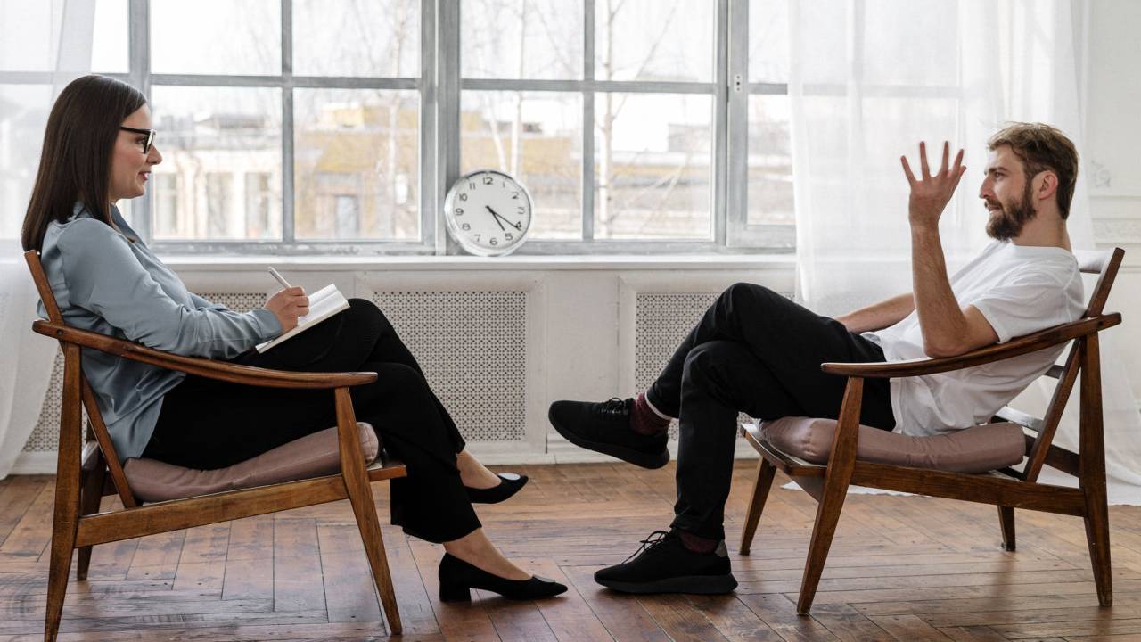 Imagem mostra uma mulher e um homem sentados frente a frente. Ele está falando e se expressando com as mãos, enquanto ela faz anotações em um caderno.