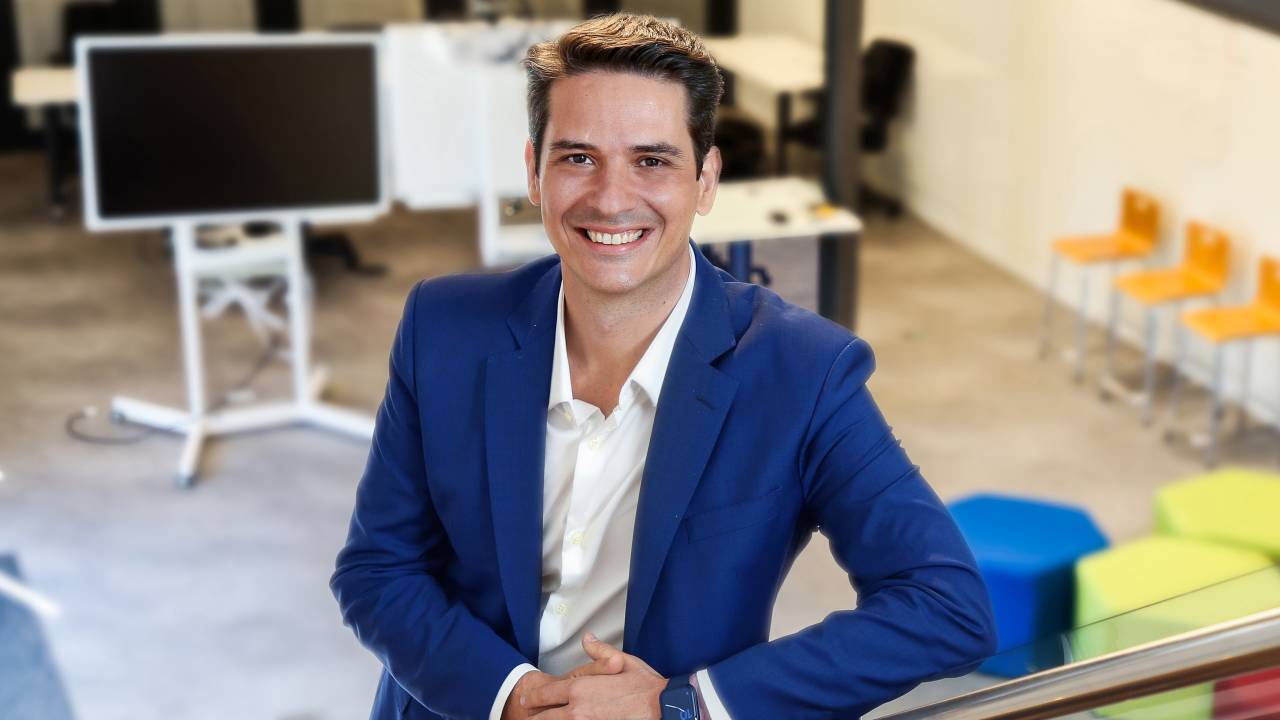 Douglas usa um blazer azul marinho por cima de uma camisa branca. Apoiado em um corrimão de um ambiente corporativo, ele sorri para a foto
