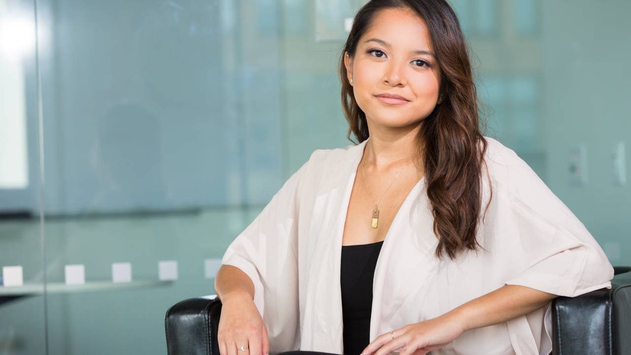 Foto mostra uma mulher morena, com sorrindo com os lábios cerrados, sentada em seu ambiente de trabalho e olhando para a câmera.