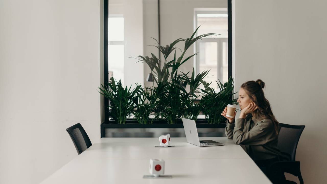 Imagem mostra uma mulher sentada em uma mesa, olhando para a tela do notebook enquanto toma café