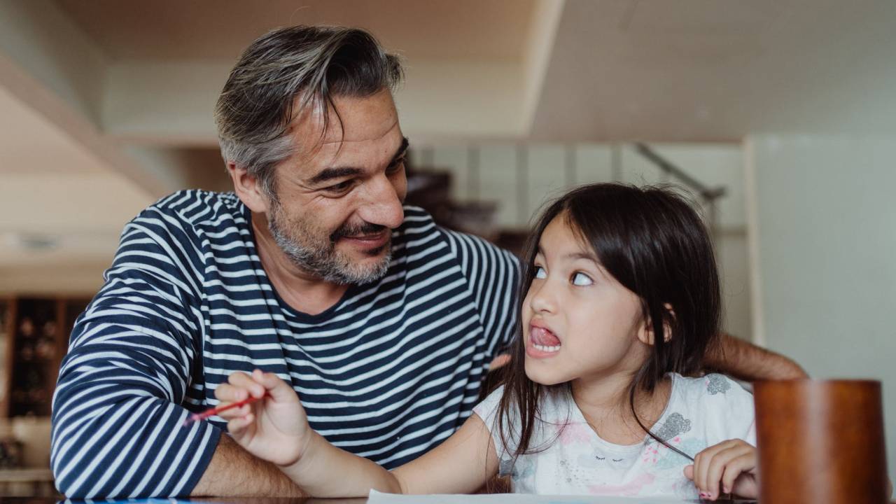 Um homem adulto está sentado ao lado de uma criança enquanto ela segura um lápis de cor e pinta uma figura