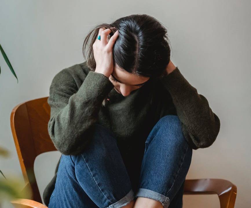 Imagem mostra uma mulher sentada em uma cadeira, aparentemente estressada. Ela segura a cabeça com as mãos e está com os pés sobre o assento