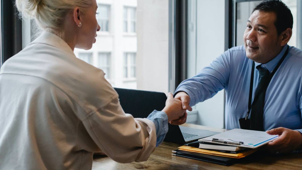Um homem vestido com uma camisa azul e gravata está sentado em frente a um computador. Do outro lado da mesa, está uma mulher. Os dois dão um aperto de mãos