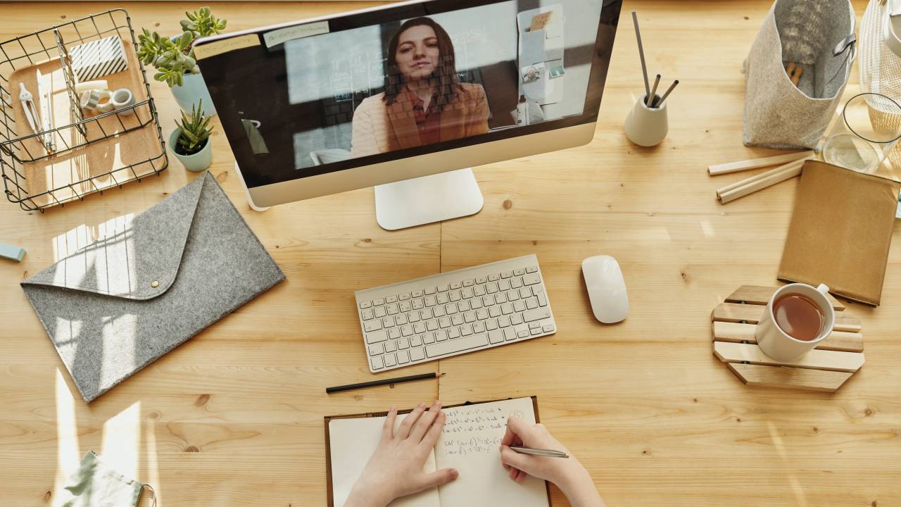 Uma pessoa faz anotações em um caderno sobre a mesa enquanto vê uma mulher dando aula pela tela do computador