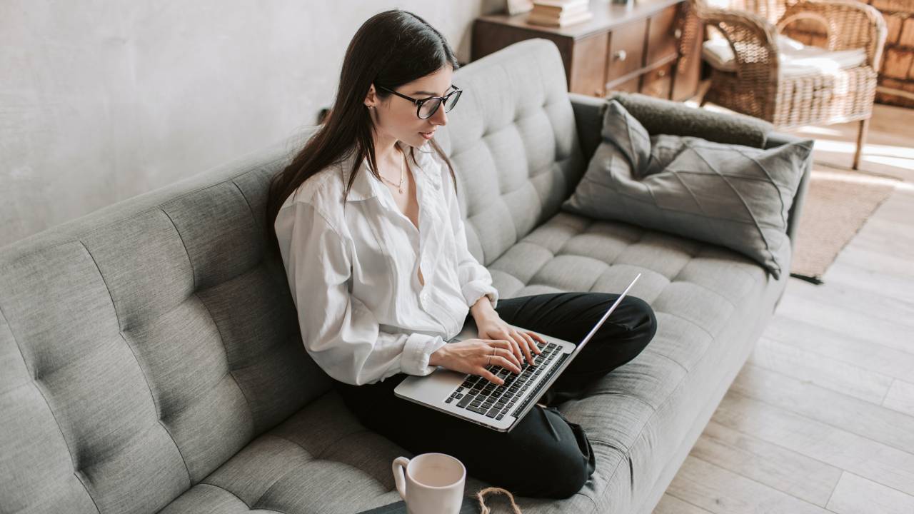 Mulher está sentada sobre um sofá cinza, digitando no teclado de um notebook. Ao lado dela há uma xícara de chá