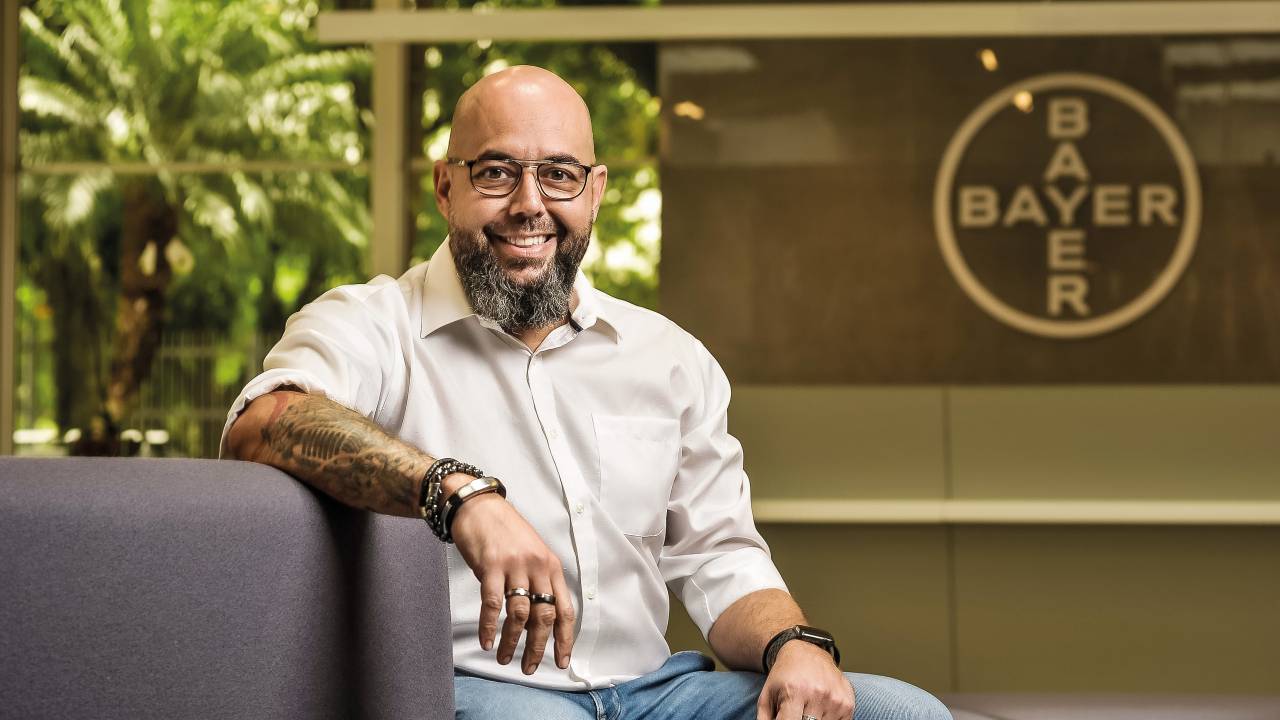 André Kraide está sentado, com o braço fechado por tatuagens apoiados no sofá. Ele veste uma camisa branca, tem barba grisalha e cabelo raspado e sorri para a foto