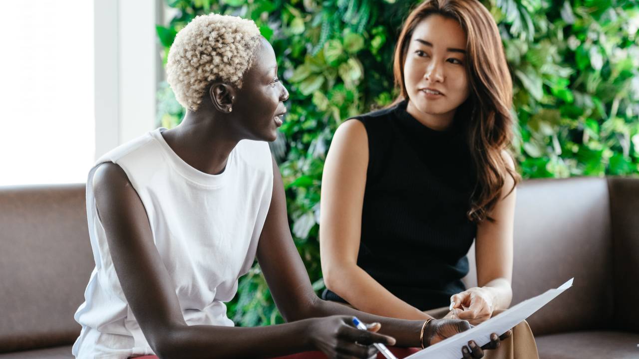 Duas mulheres conversam entre si. As duas estão sentadas. Uma delas é negra e tem os cabelos curtos loiros bem claros, a segunda tem traços orientais e é morena.