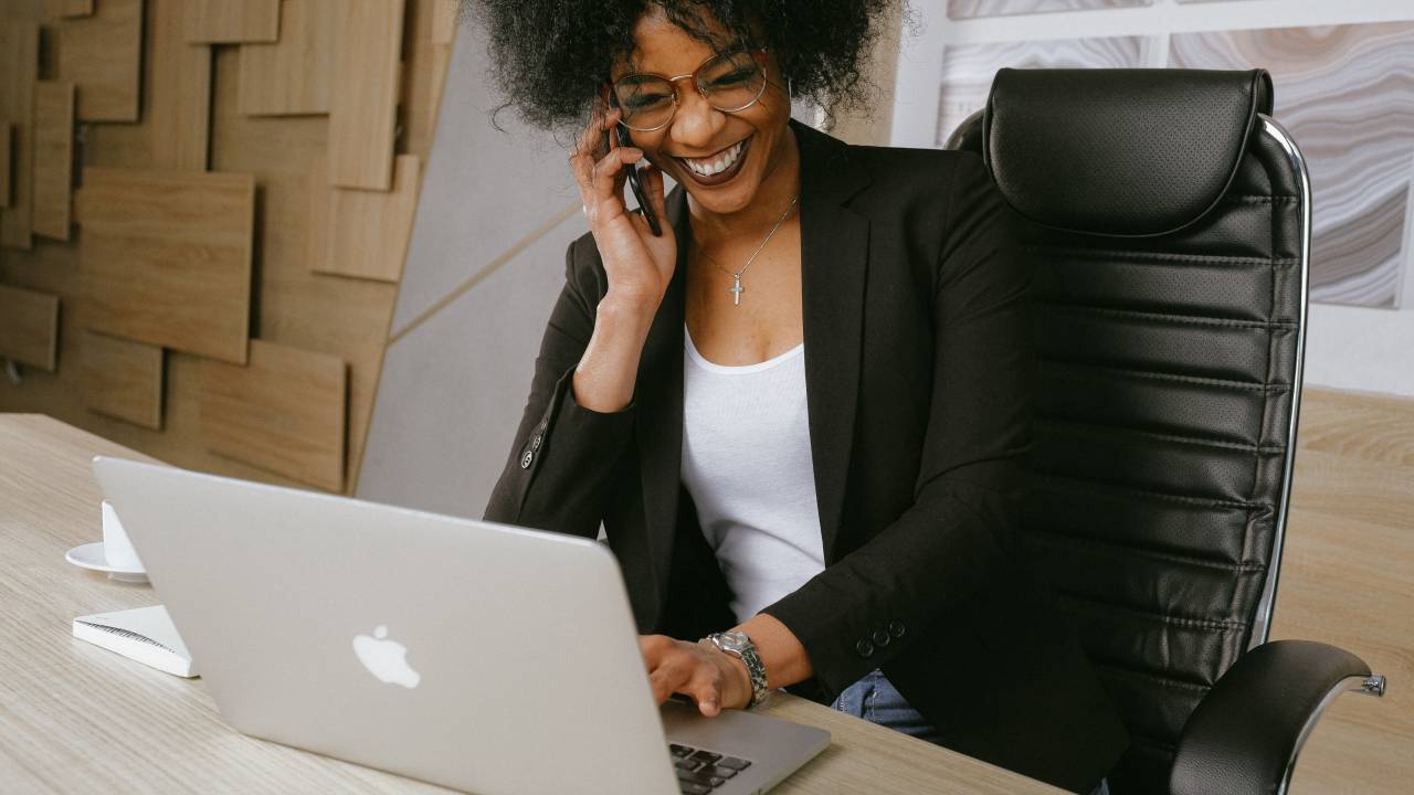Mulher negra fala ao celular enquanto digita no notebook. Ela está sorrindo