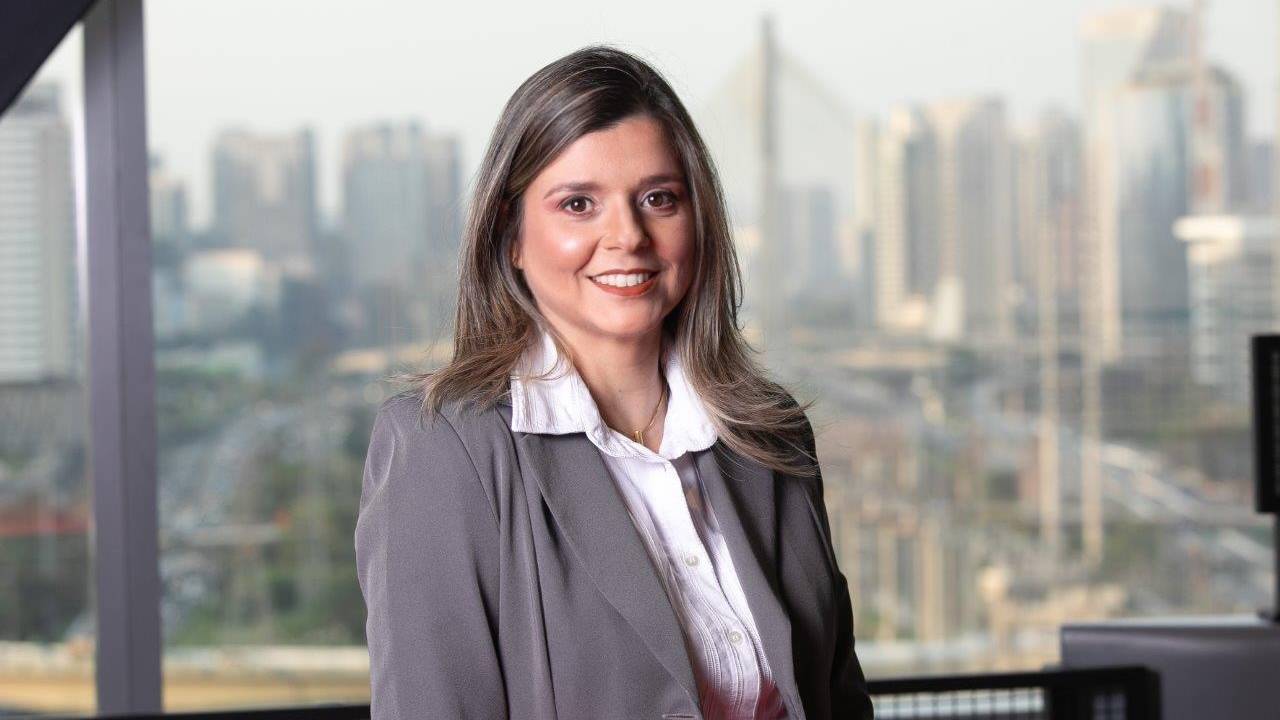 Karine está vestindo uma camisa branca e, por cima, um blazer cinza escuro. Ela está sorrindo e posando em frente a uma janela grande cuja vista é a ponte estaiada de São Paulo