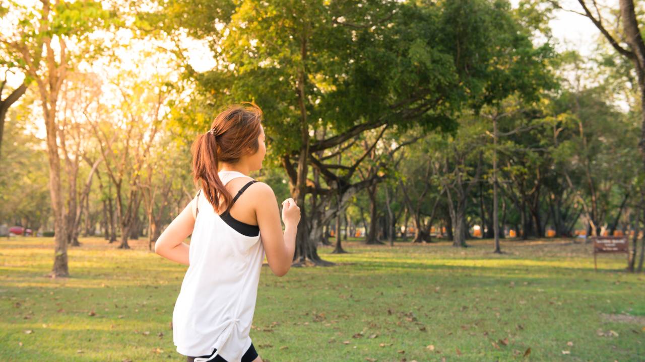 Uma mulher está correndo em um parque. Ela veste uma camiseta branca, um short preto e está com o cabelo preso em um rabo de cavalo. O parque tem grama e muitas árvores