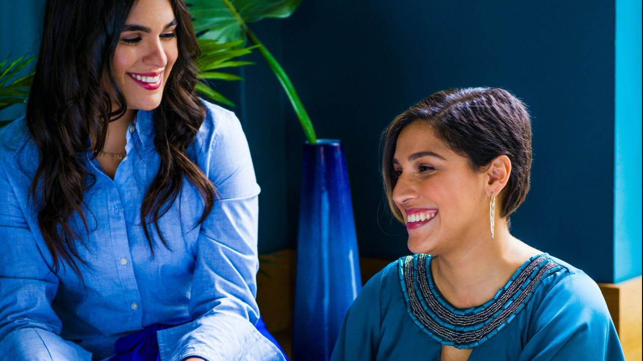 Duas mulheres conversam entre si. As duas estão sentadas e vestem tons entre azul e verde. Atrás delas, há um vaso com uma planta. Elas estão em um ambiente fechado.