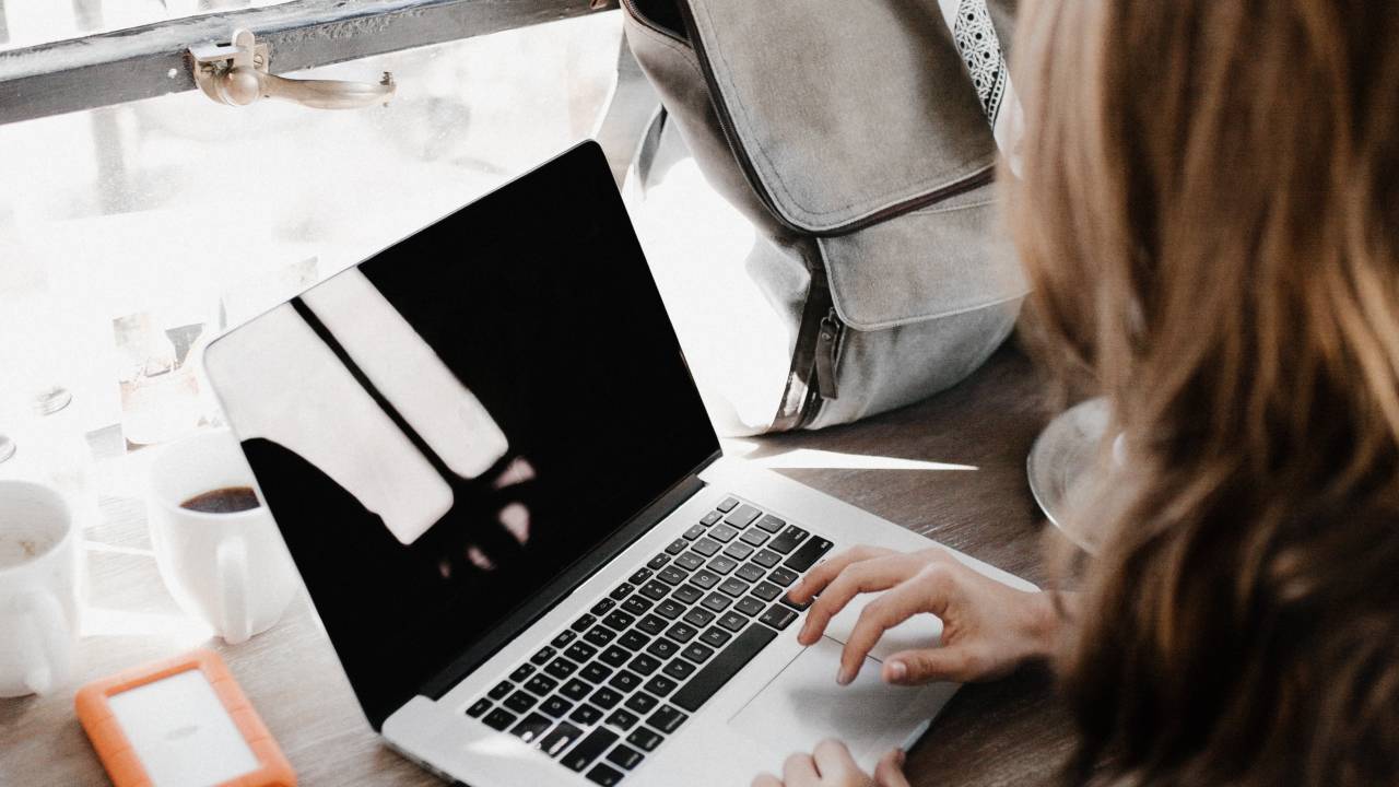 Uma mulher está trabalhando em um notebook. Sua mochila está na mesa, ao lado do computador.