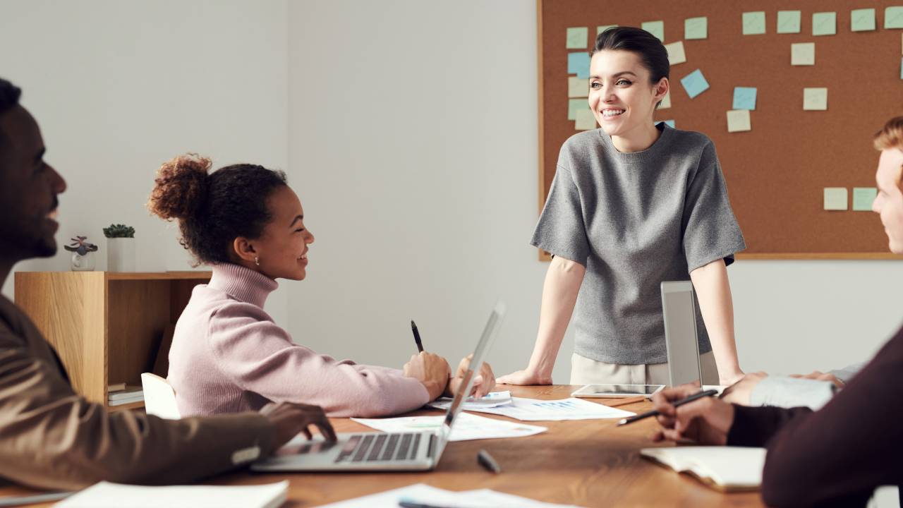 Uma mulher está de pé apoiada com os braços em uma mesa. Ela parece liderar alguma reunião. A mesa está cheia de materiais de trabalho, como canetas, computador e folhas.