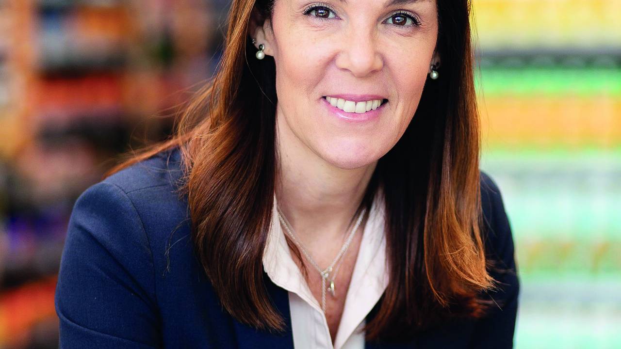 Catia posa com um blazer azul marinho, uma camisa branca, em um fundo desfocado cheio de gôndolas de mercadorias