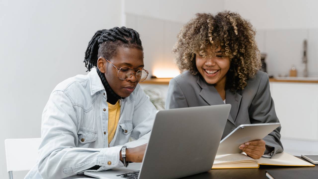 Dois profissionais negros estão sorrindo enquanto olham para a tela de um computador. Os dois estão sentados em uma mesa de escritório