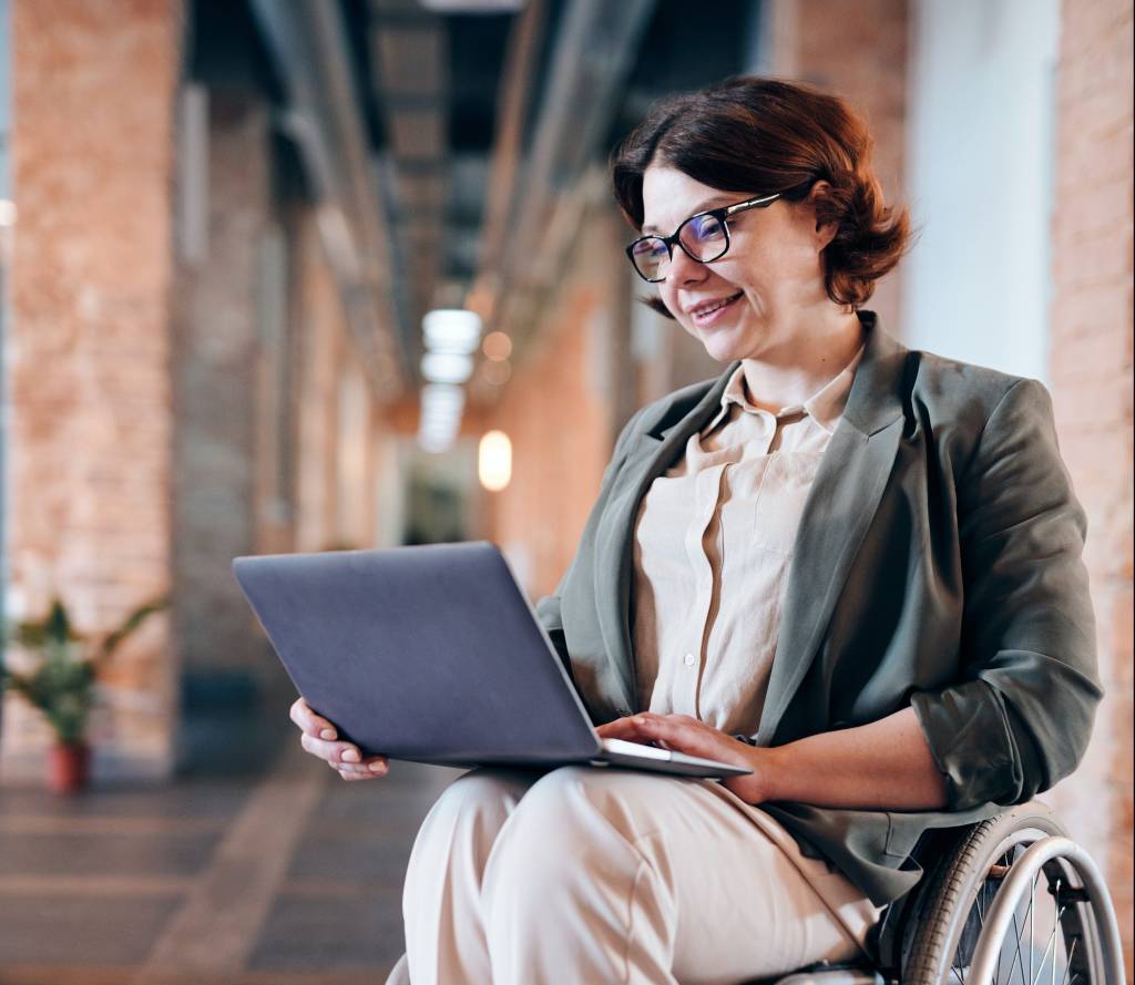 Uma mulher está sentada sobre uma cadeira de rodas e tem um computador apoiado no colo. Ela interage com a tela do computador enquanto sorri