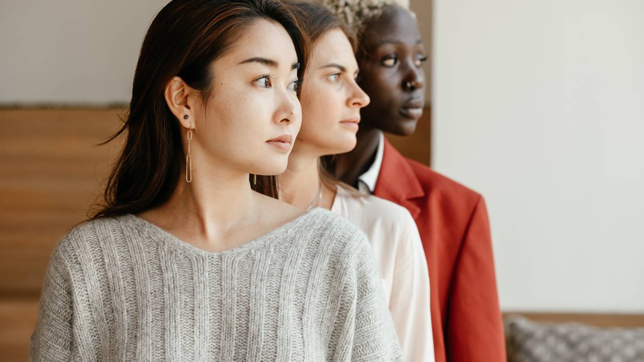 Três mulheres estão uma na frente da outra. Elas olham para o lado. A primeira tem traços orientais, a segunda é branca de cabelos escuros e a terceira é negra de cabelo curto e loiro.