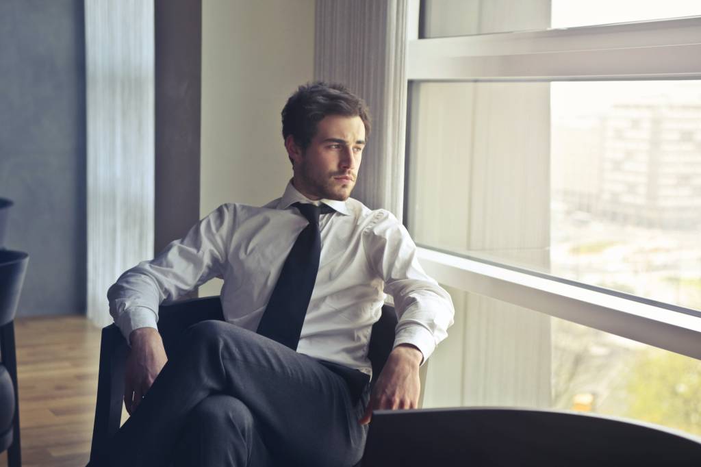 Homem de camisa, gravata e calça social olha pensativo pela janela