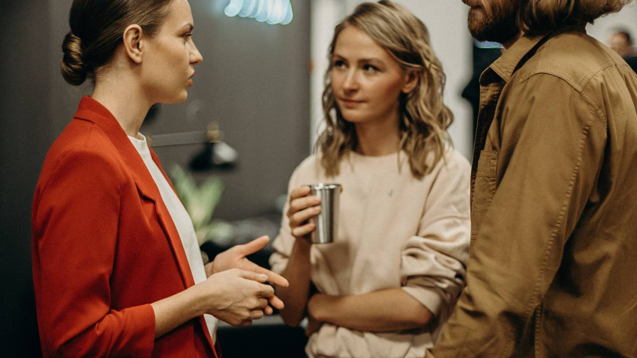 Três pessoas estão de pé conversando. Todas usam roupas de frio, uma veste vermelho, a outra rosa claro e a terceira marrom. Uma delas está com uma bebida na mão