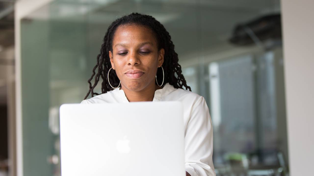 Uma mulher negra está atrás de um computador. Ela está com os lábios cerrados, vestida de branco, prestando atenção na tela