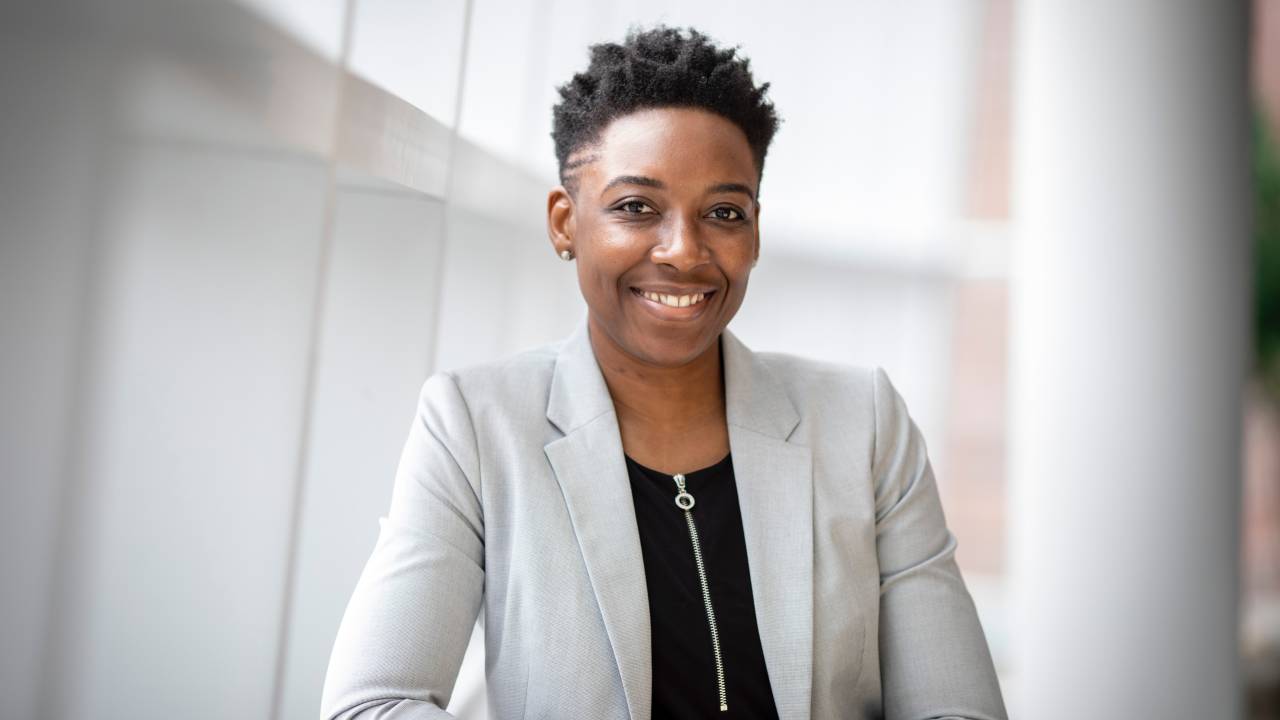 Uma mulher negra e de cabelos curtos sorri. Ela usa um blazer cinza e uma blusa preta por baixo e está em um fundo neutro