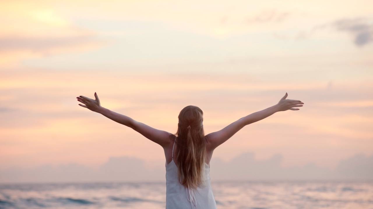 Uma mulher está de costas para a foto, mas de frente para o mar. Ela está de cabelos soltos, braços abertos e com um vestido branco