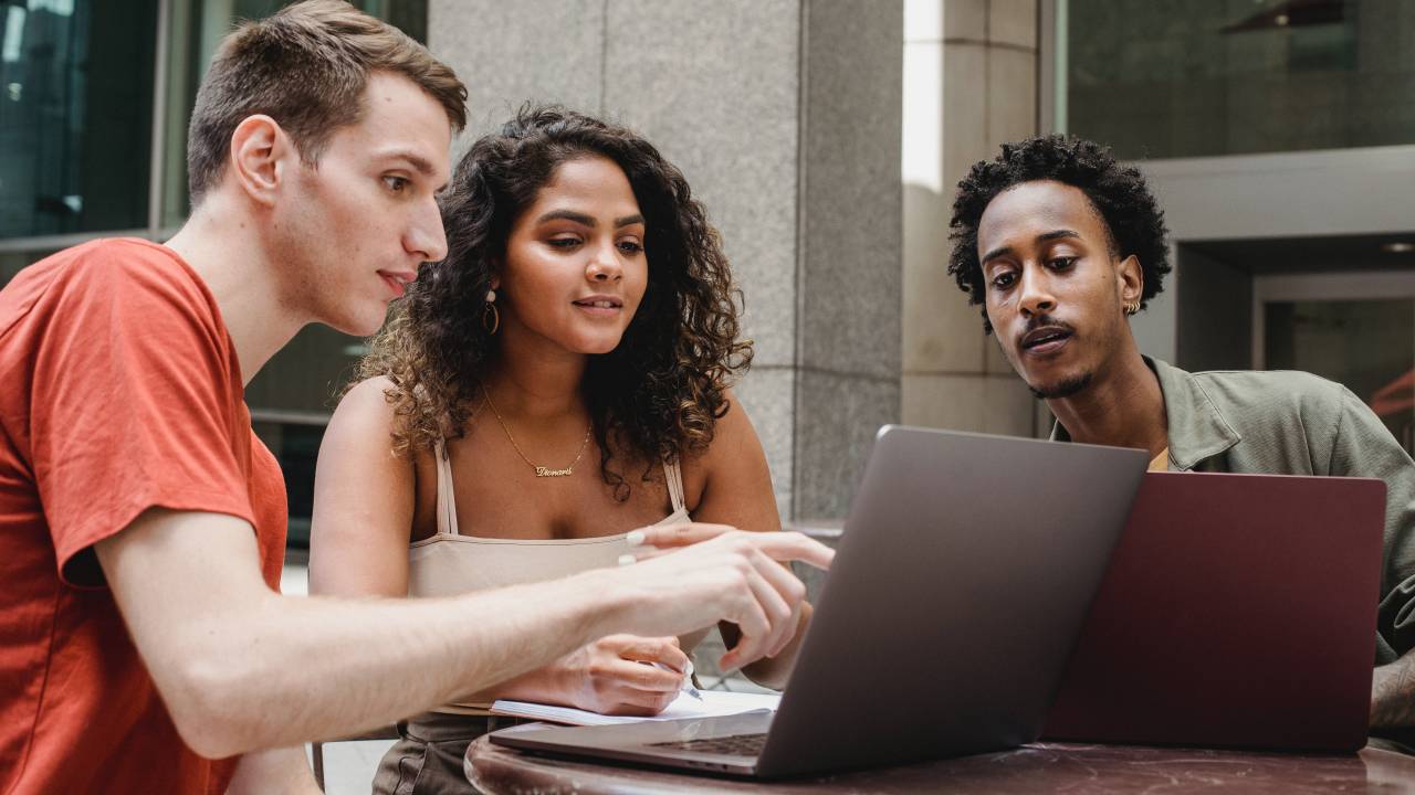 Três pessoas estão sentadas ao redor de uma mesa, ao ar livre, enquanto observam a tela de um computador