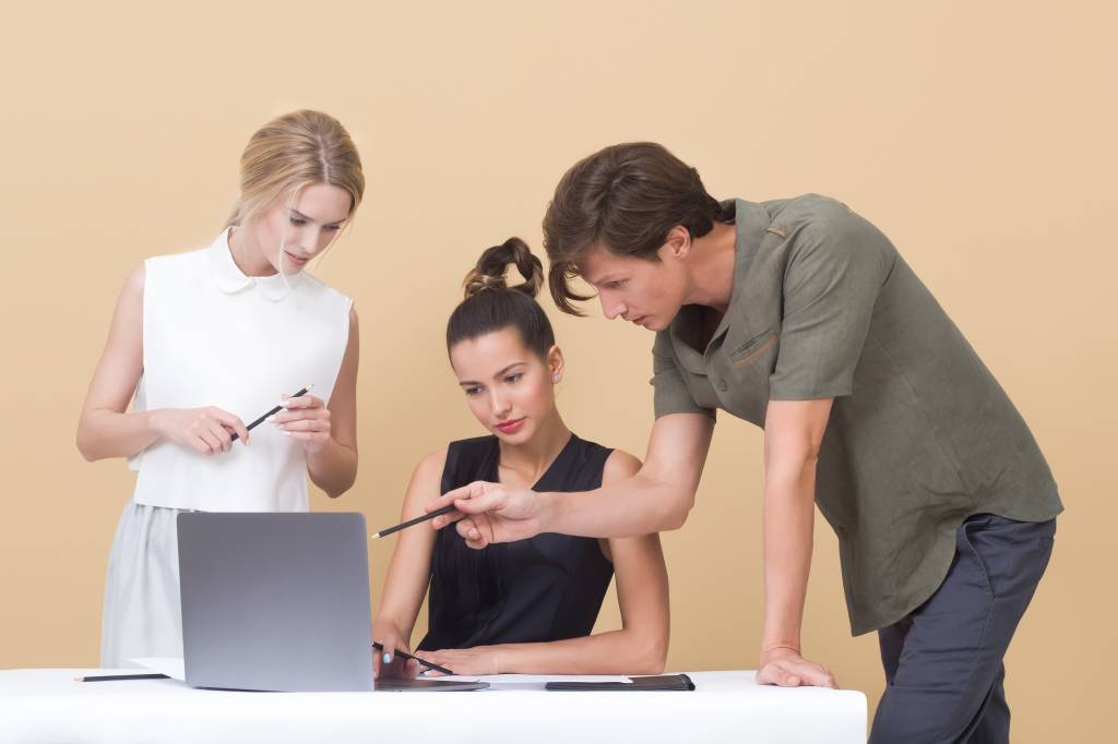 Três pessoas, uma delas sentada, estão analisando a tela de um computador. Os três têm um lápis em mãos e estão em frente a um fundo de cor sólida
