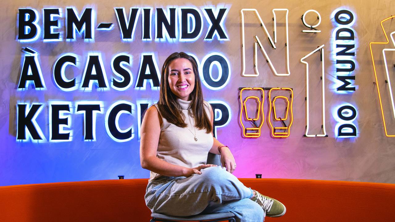 Carolina está sentada em um sofá vermelho aveludado, atrás dela há um letreiro em neon escrito: "Bem-vindo à casa do ketchup"