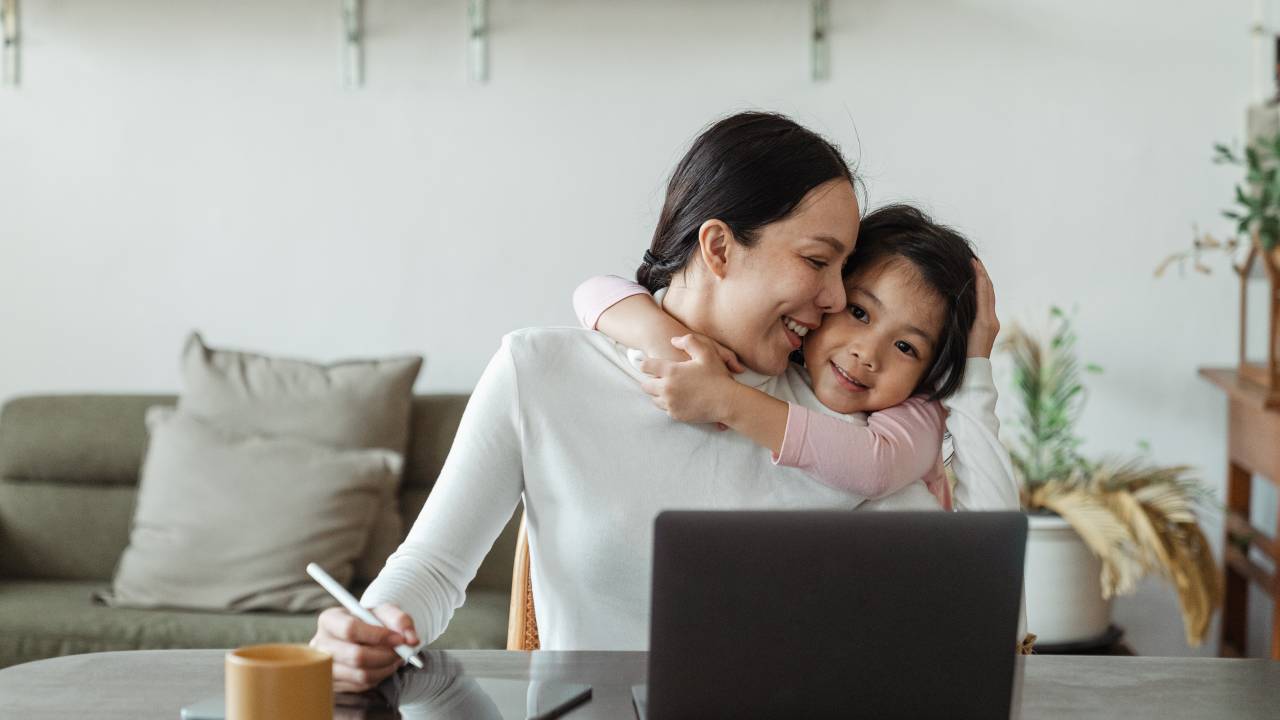 Uma mulher está sentada, em frente a um computador. Ela está sendo abraçada por sua filha, que é criança