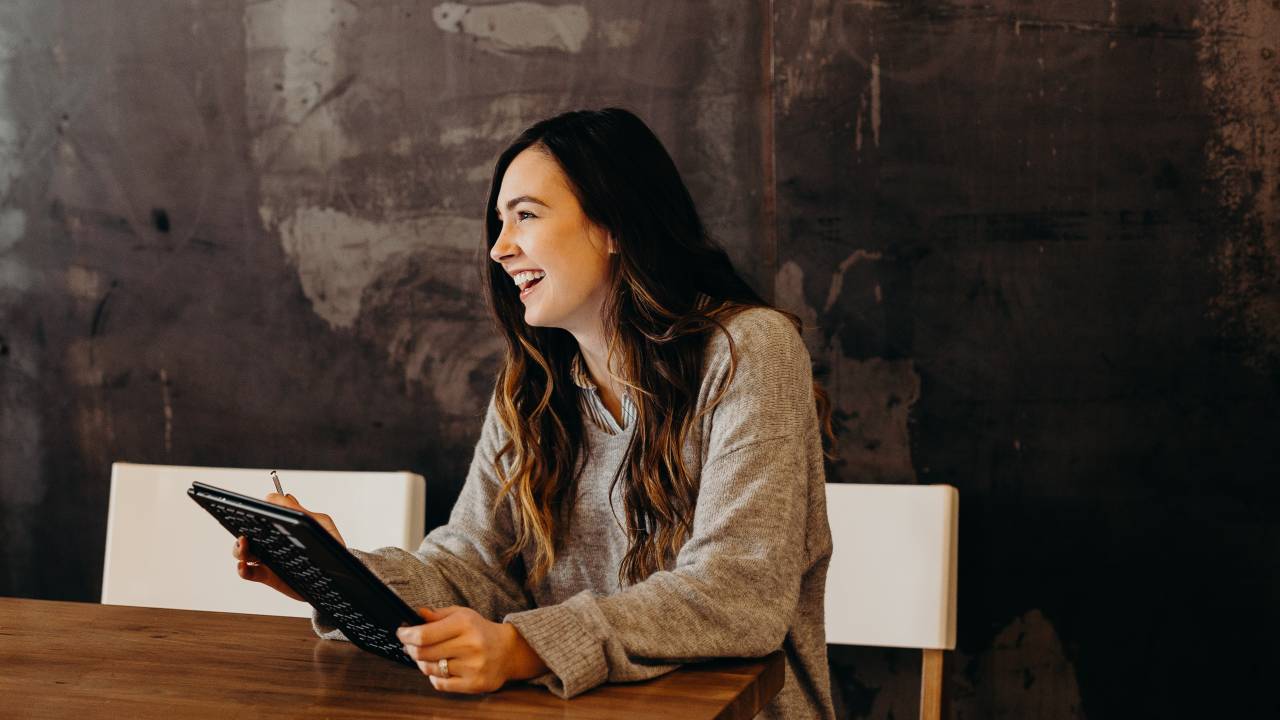 Uma mulher está sentada com os braços apoiados em uma mesa de madeira. Ela está sorrindo e tem um tablet nas mãos