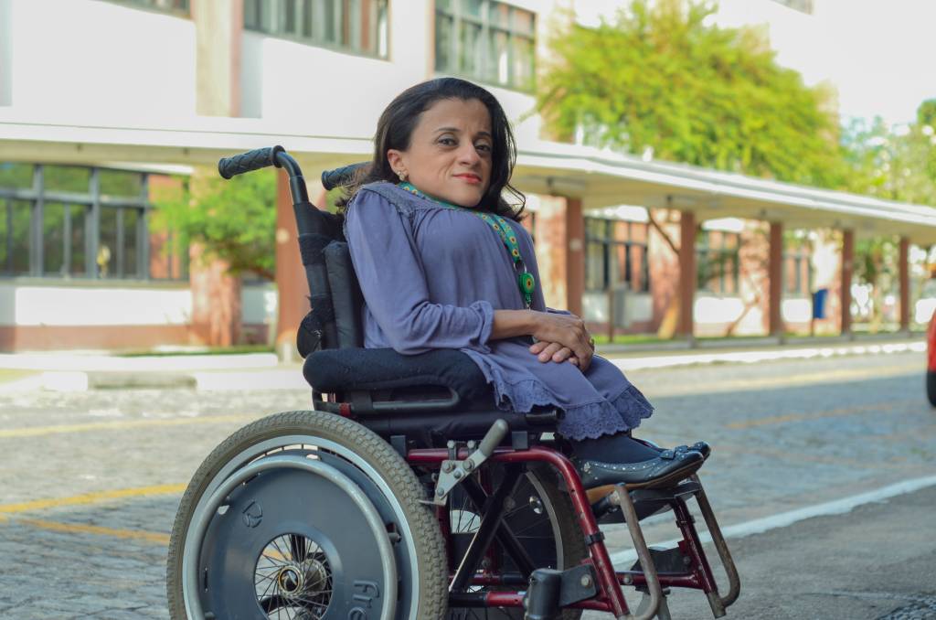 Thais Pessanha está sentada em uma cadeira de rodas. Ela está de lado, olhando para a foto, vestindo roupas em tom de azul em um ambiente externo