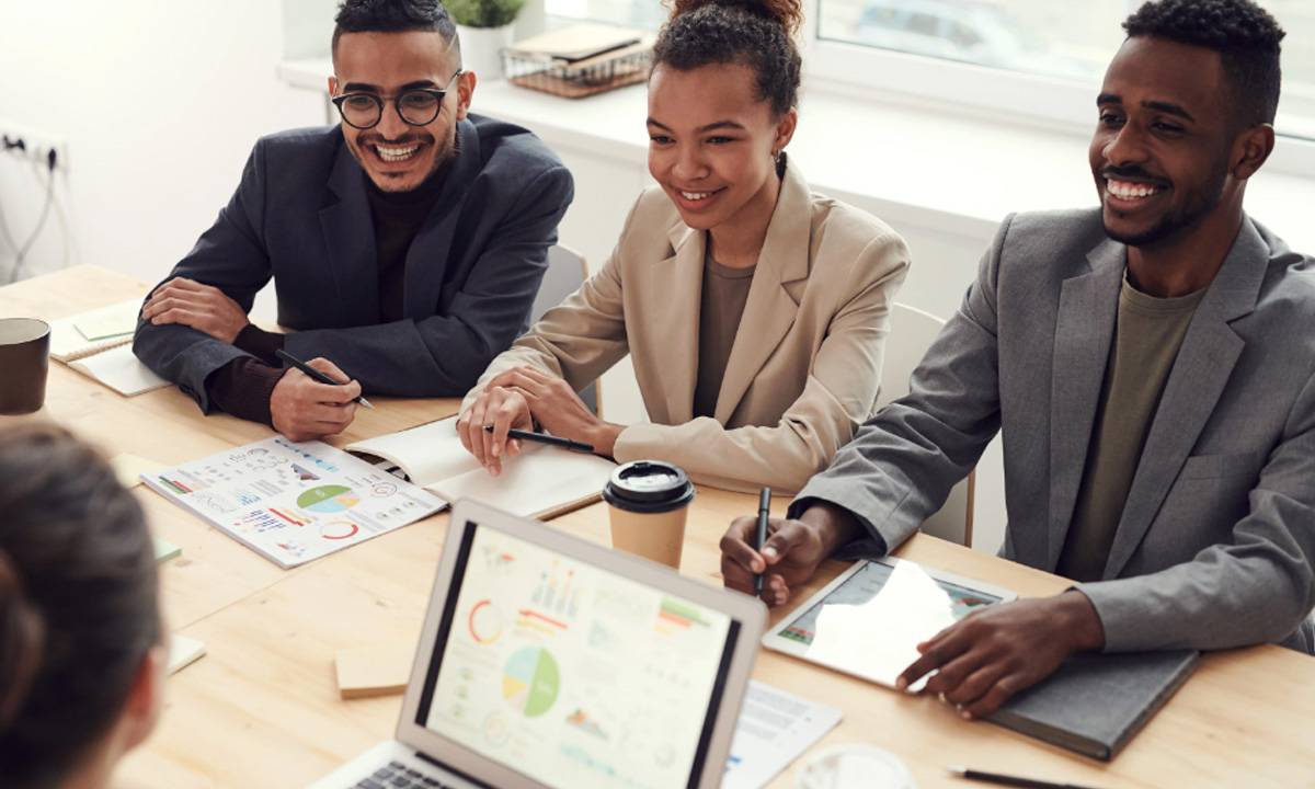 Três funcionários sorriem enquanto conversam com uma quarta pessoa, que está do outro lado da mesa