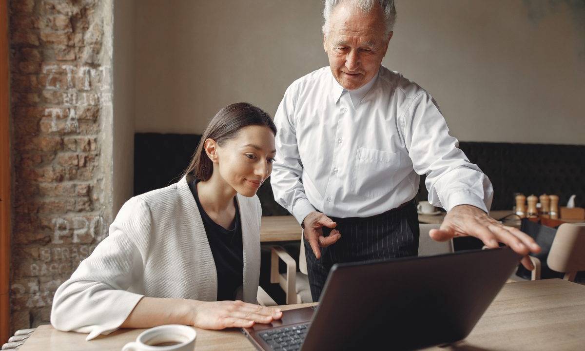 imagem de dois colegas de trabalho, um homem idoso e grisalho ao lado de uma mulher jovem e branca.