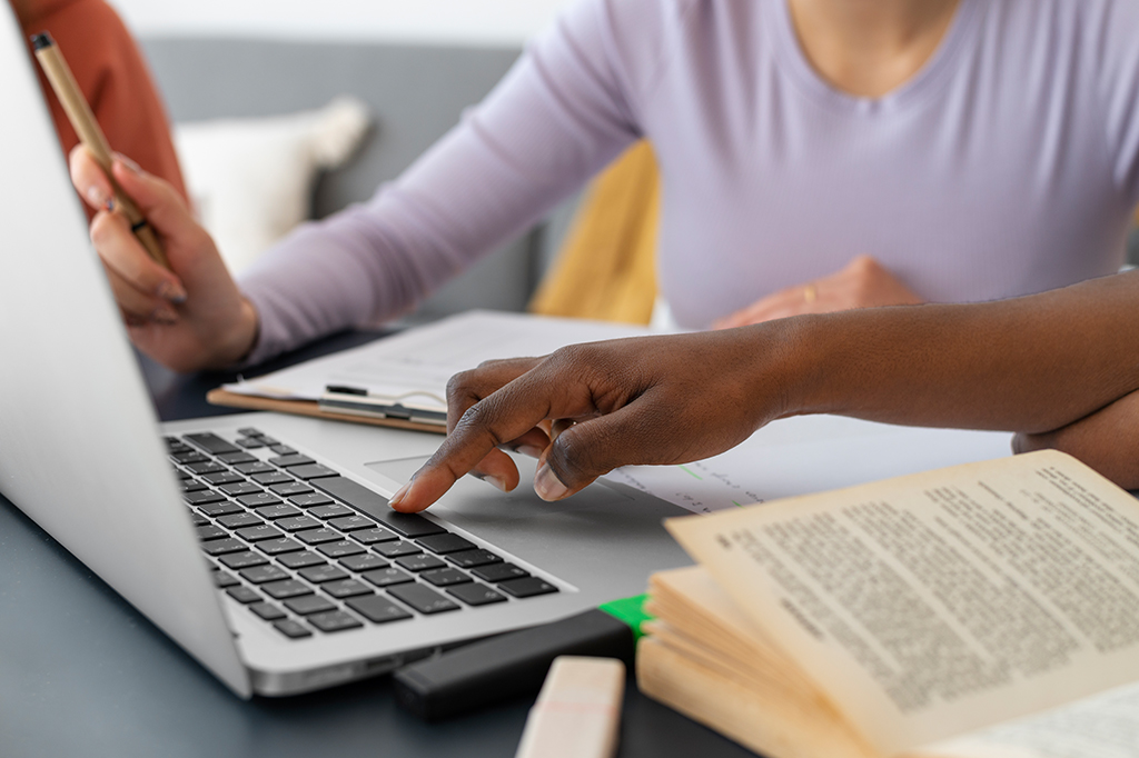Duas pessoas sentadas em uma mesa. Há um livro, papéis e um notebook sobre ela.