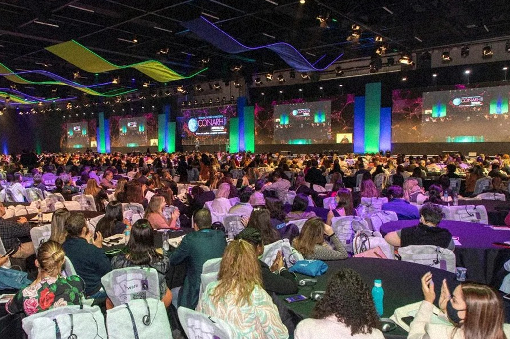 Um evento com diversas pessoas sentadas em mesas coletivas diante de um palco.